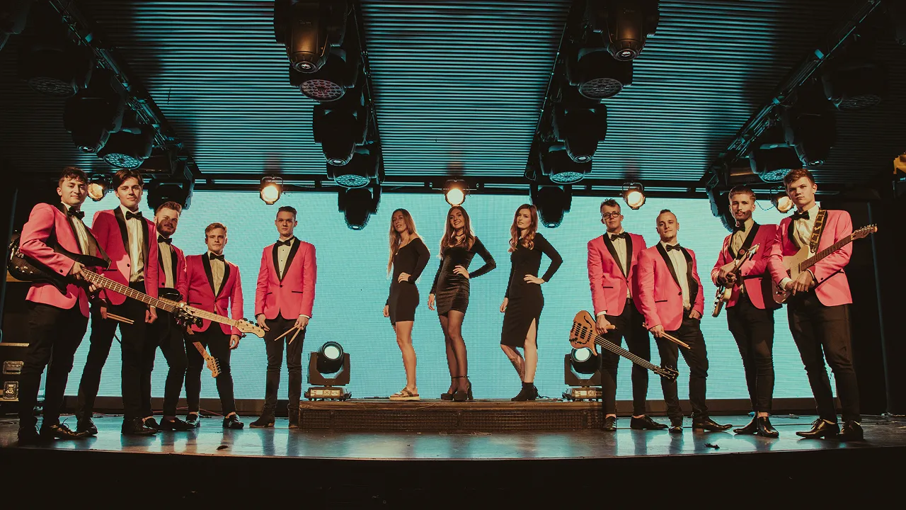 A band on stage with eight men in pink jackets holding instruments and three women in black dresses standing on a raised platform.