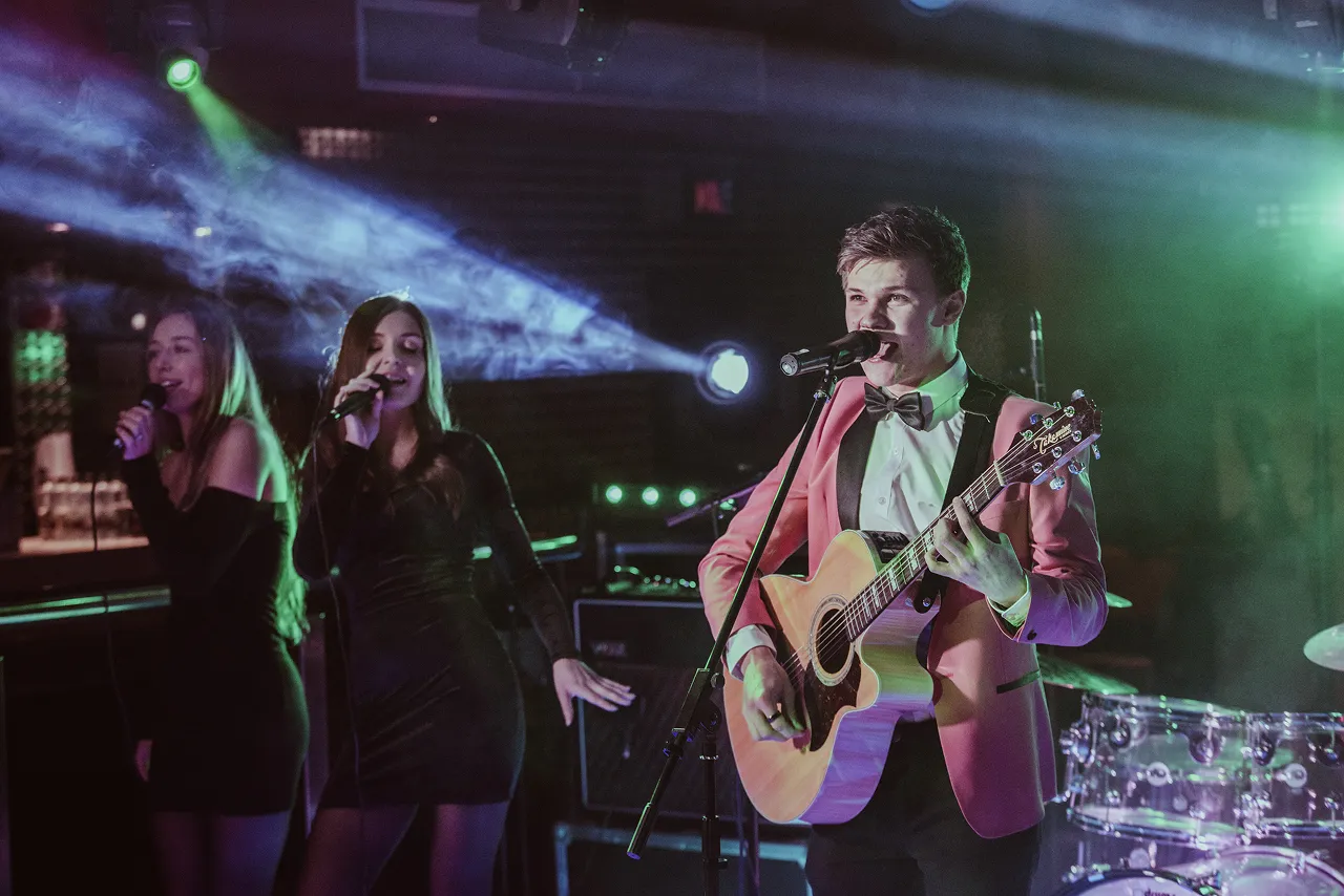 Male musician in a pink jacket playing an acoustic guitar and singing into a microphone with two female vocalists in black dresses singing beside him on stage.
