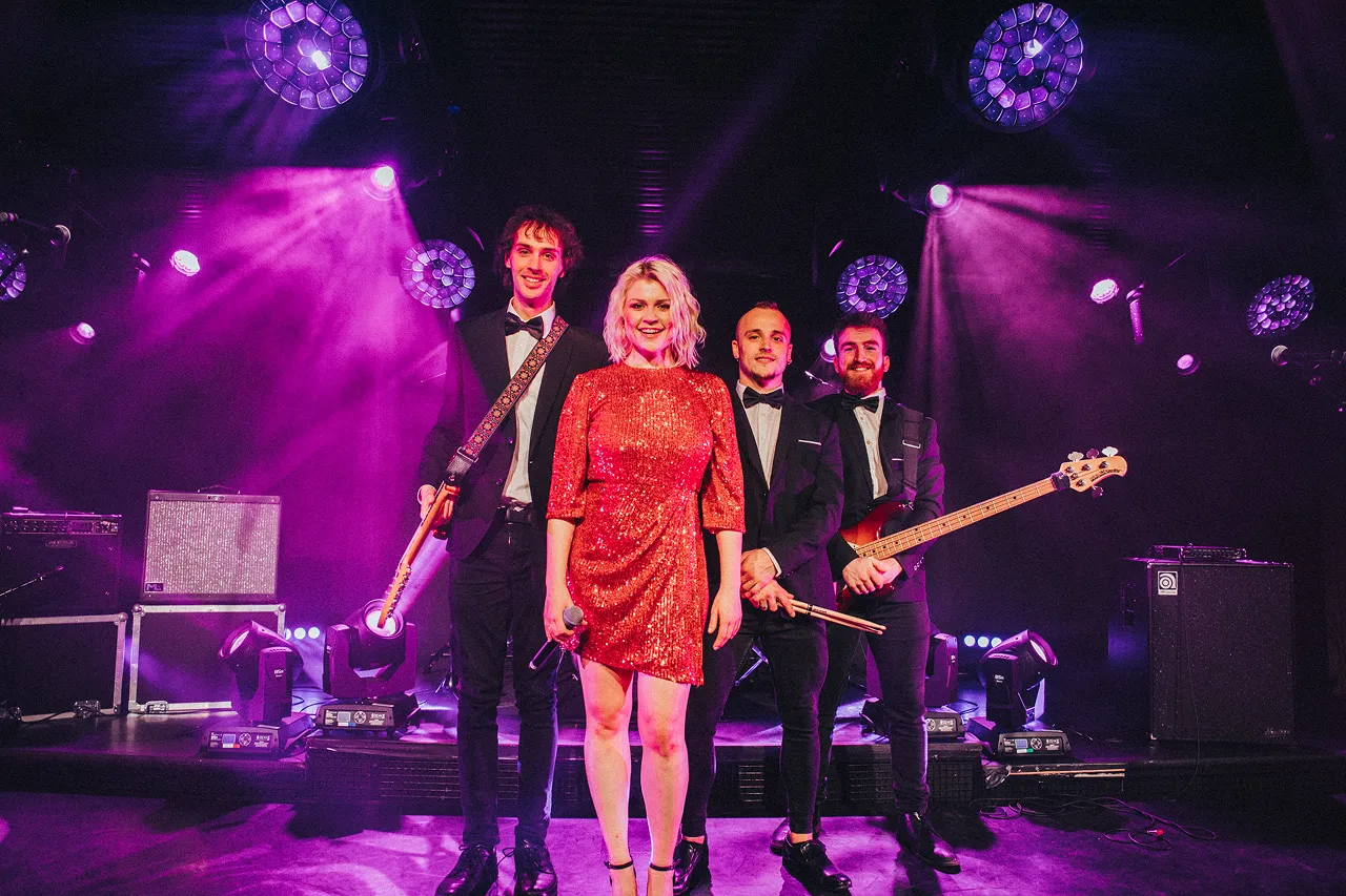 Four musicians wearing formal attire on stage with purple lighting, including a woman in a red sequined dress holding a microphone and three men holding instruments.