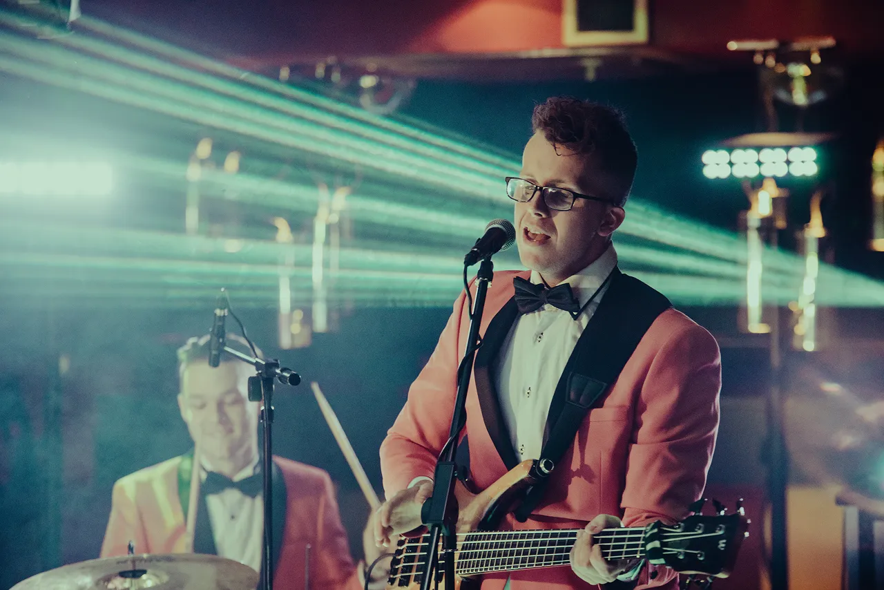 Male musician in a pink suit with black lapels singing and playing electric bass guitar with drummer in the background under blue stage lights.