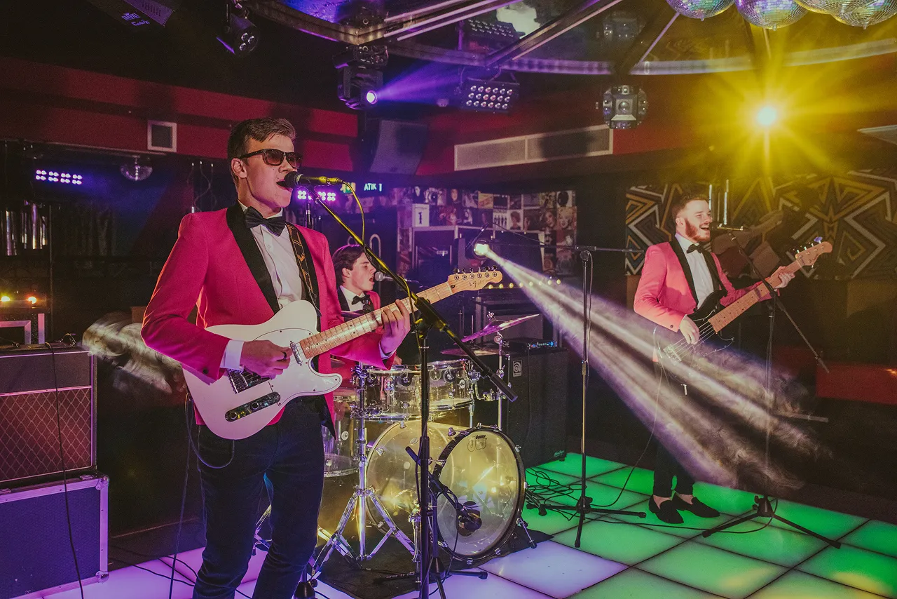 Three-member band performing live on a lit dance floor with colorful stage lights in a club setting.