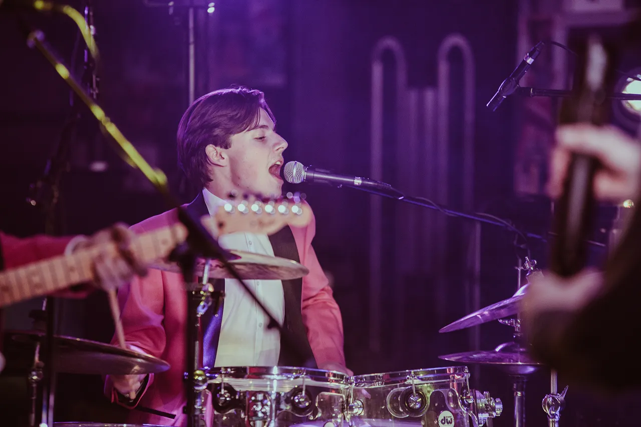Young male drummer and vocalist singing into a microphone while playing a clear drum set on stage.