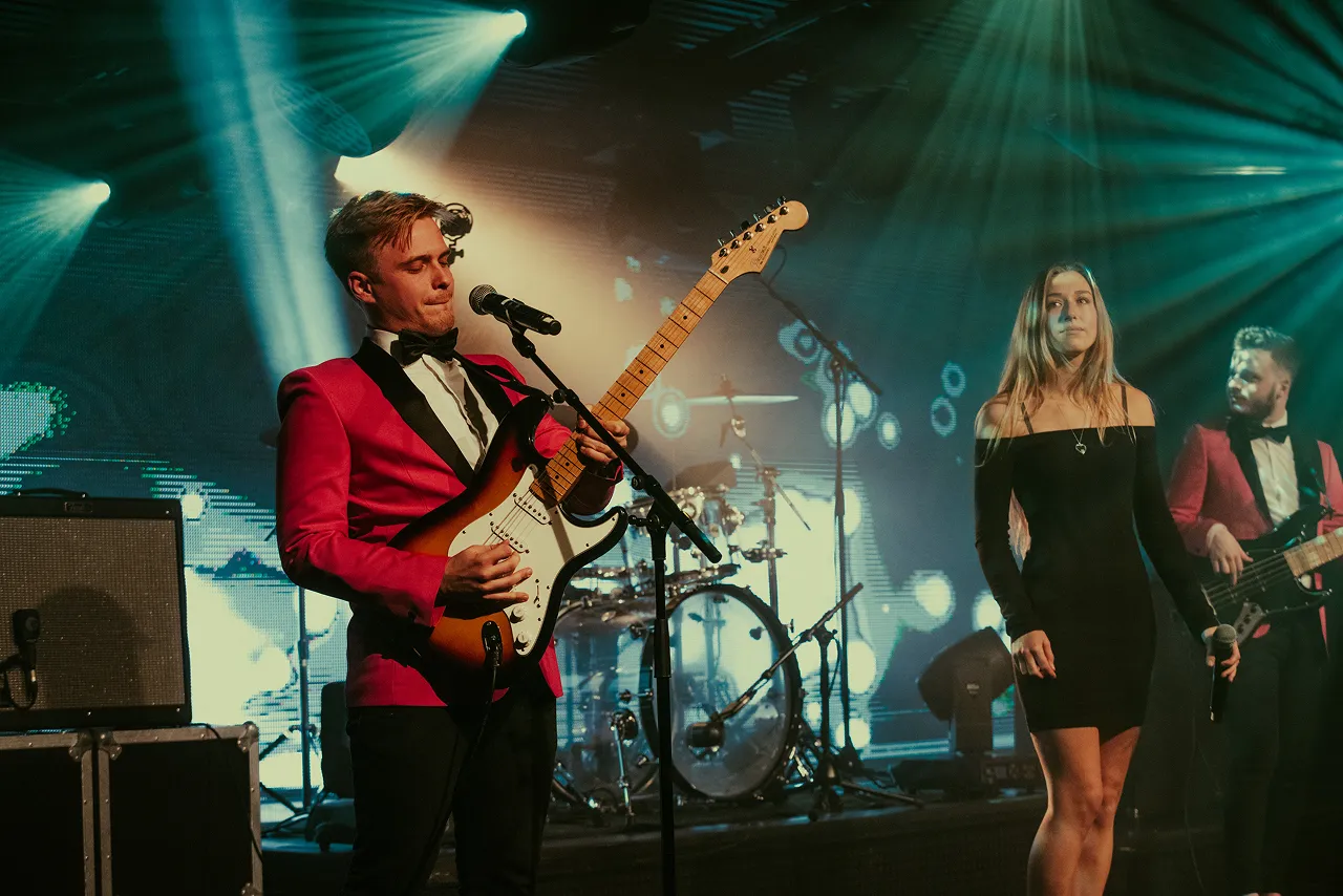 Band performing on stage with a guitarist in a red jacket playing electric guitar, a woman in a black dress holding a microphone, and a bassist in a red jacket under blue stage lights.