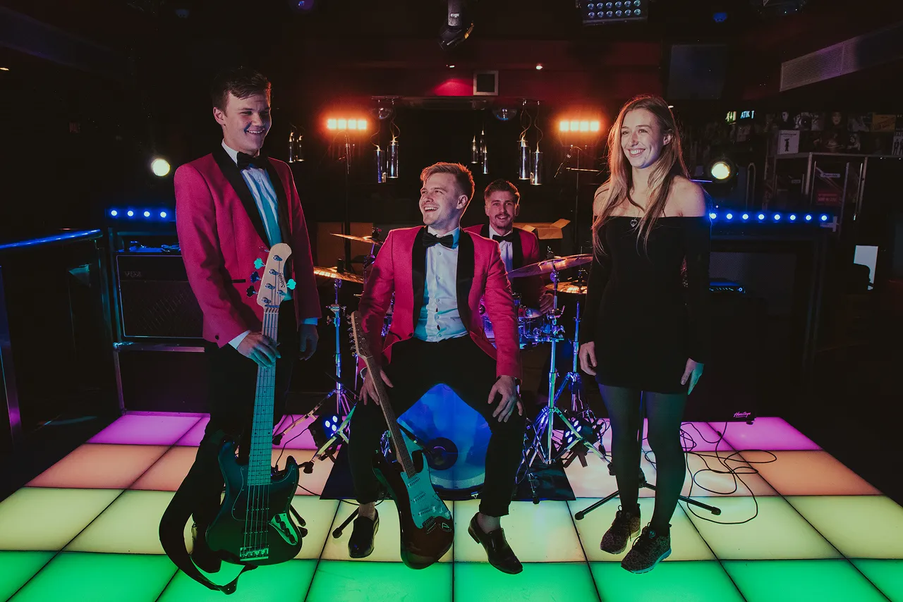 Four band members in formal attire on a colorful lighted dance floor, two holding guitars and one sitting behind a drum set.