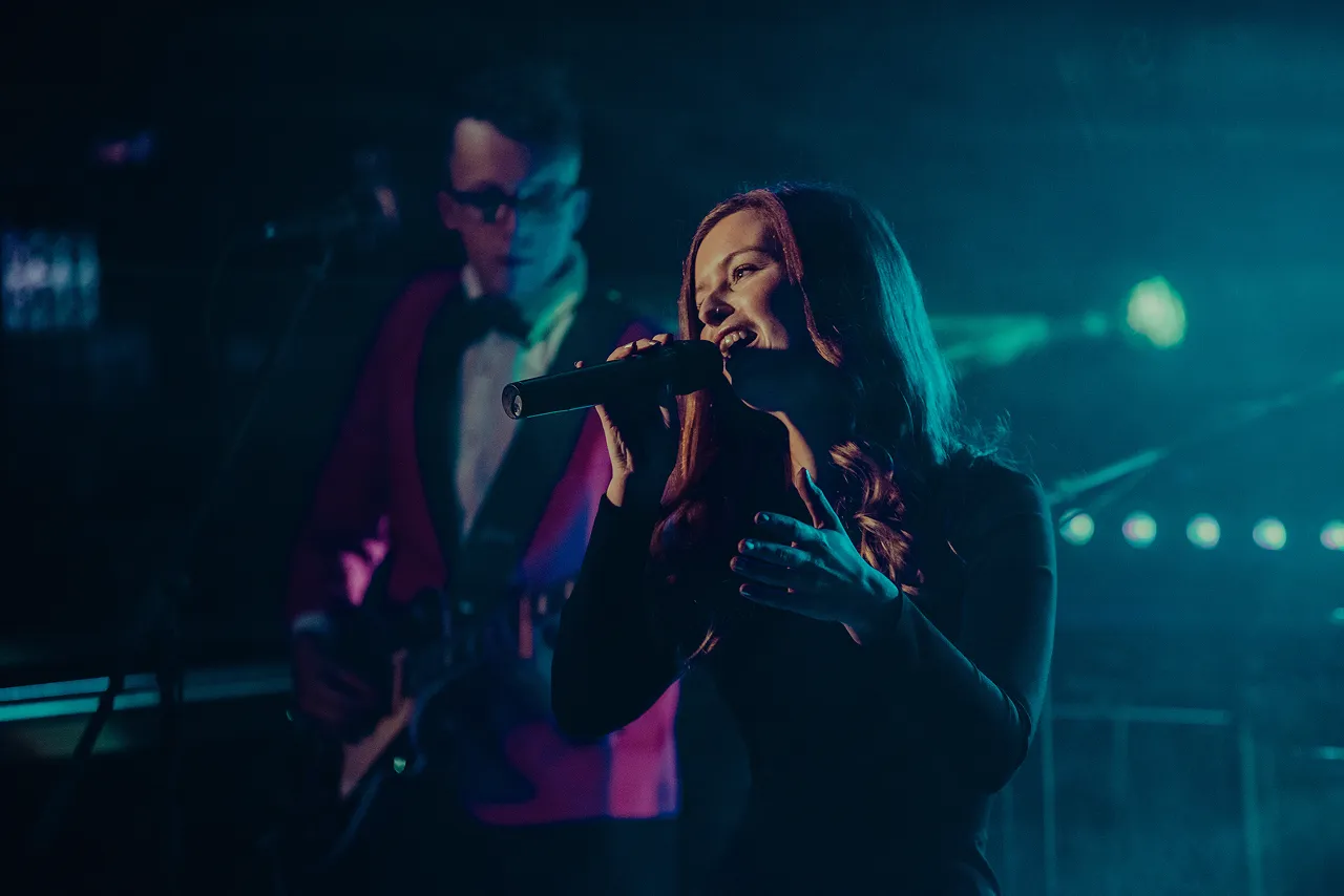 Female singer with long hair holding a microphone and performing on stage with a guitarist in the background under dim blue lighting.