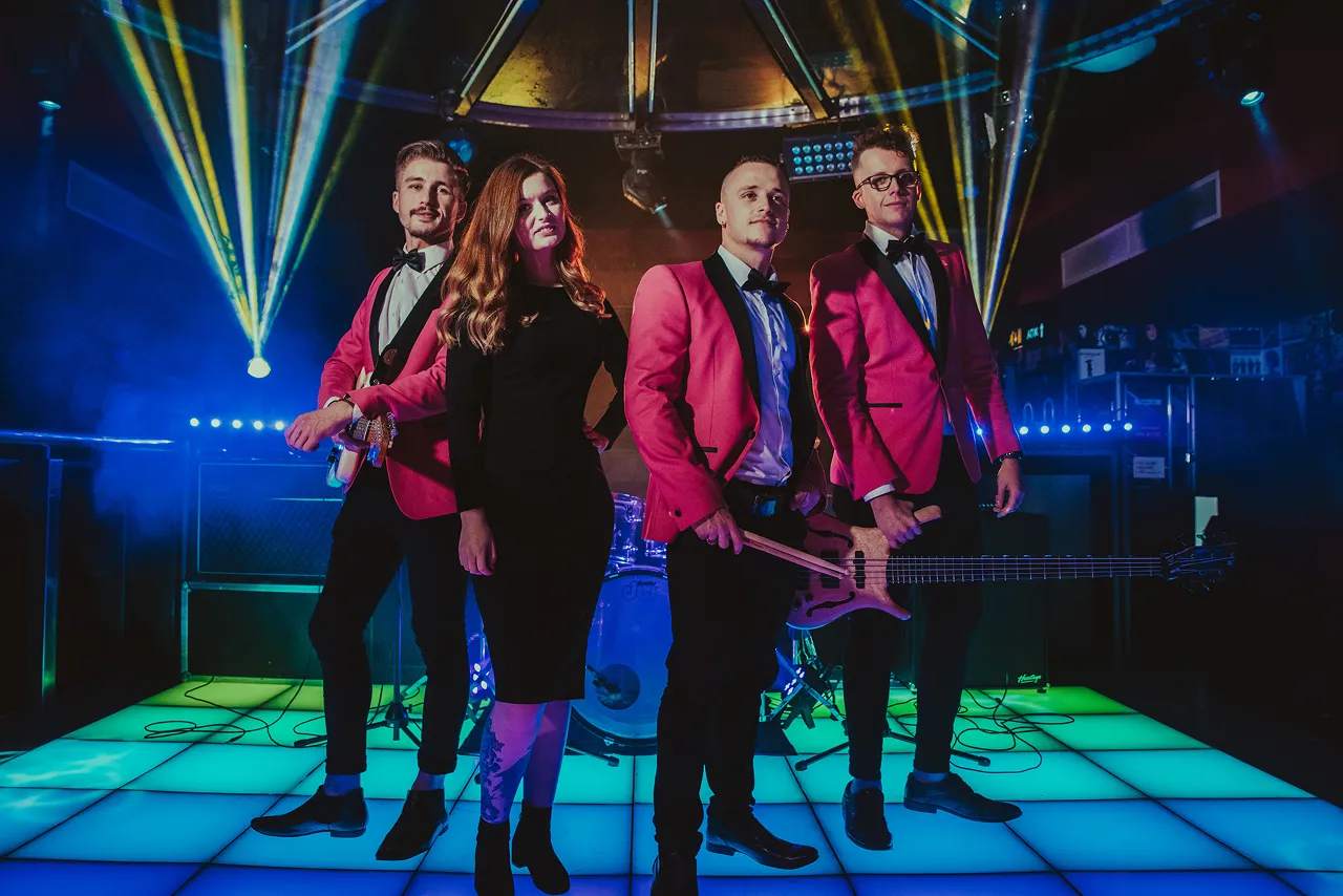 Four musicians standing on a brightly lit stage with blue and green floor panels and colorful spotlights behind them.