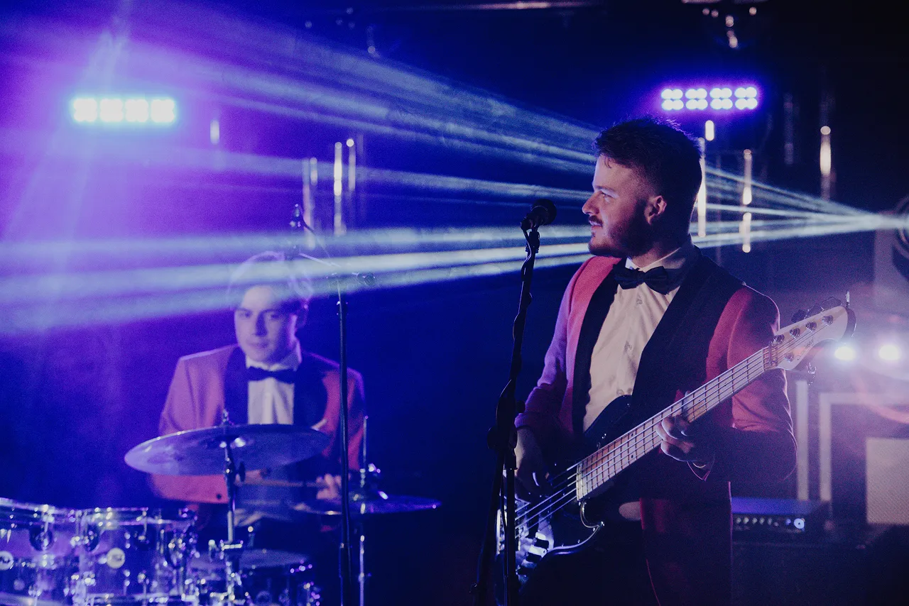 Two musicians dressed in matching red jackets with black lapels and bow ties performing on stage, one playing drums and the other playing bass guitar under blue and purple stage lights.