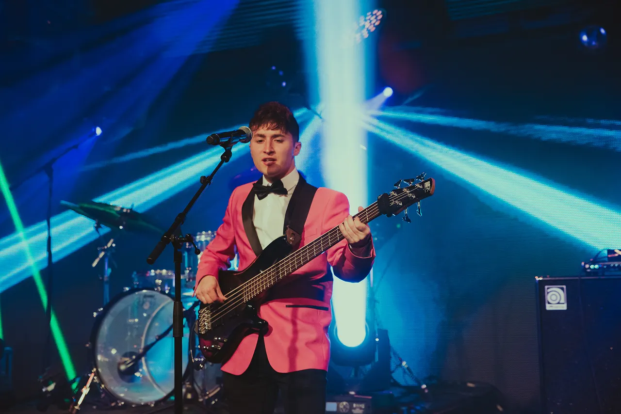 Male musician in a pink tuxedo jacket playing bass guitar and singing on stage with blue and green stage lights.