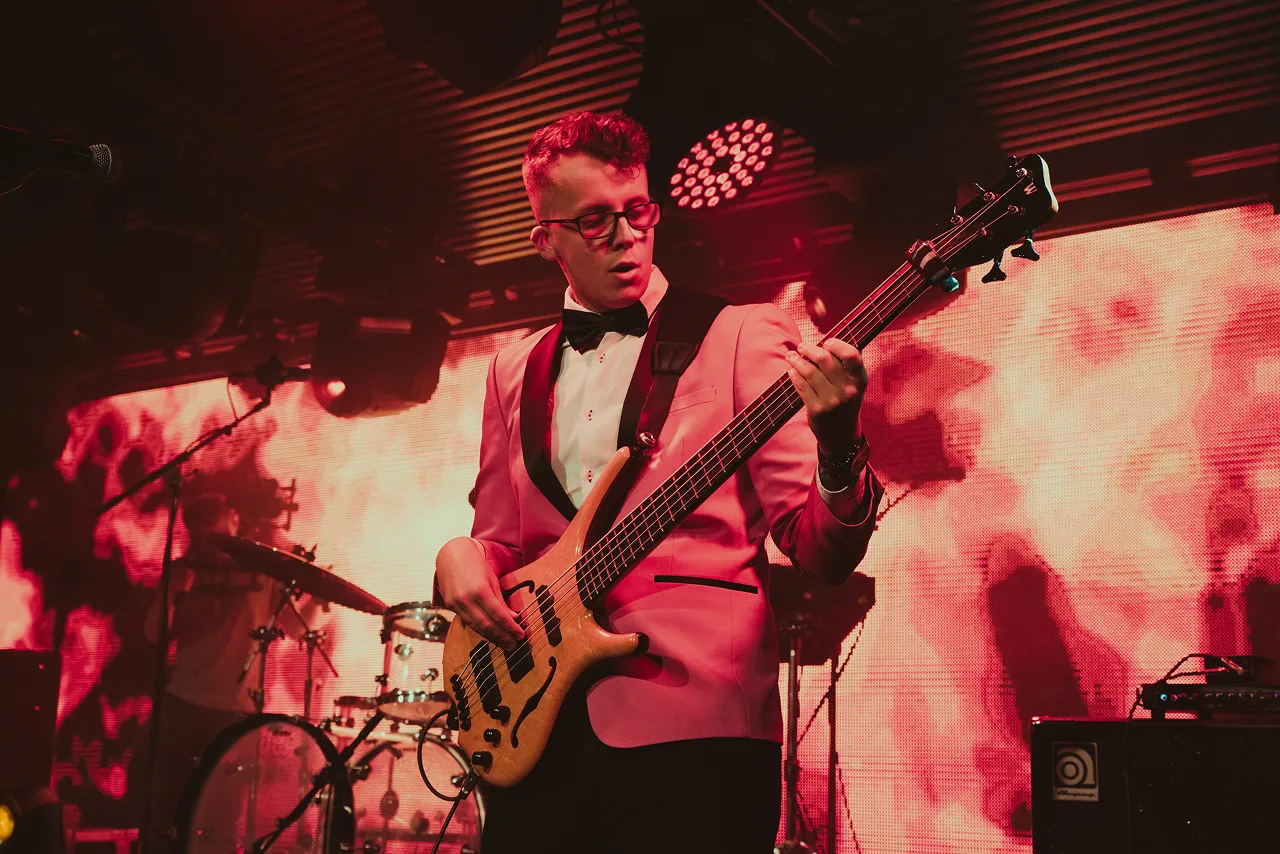 Musician in a red and black tuxedo playing a bass guitar on stage with red lighting and drums in the background.