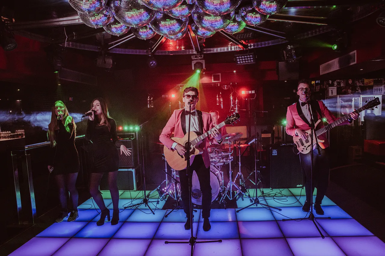 Band performing on a glowing blue tiled stage under multiple disco balls with two female singers, a guitarist, a bassist, and a drummer.