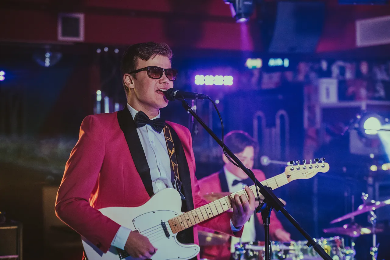Man in a red tuxedo jacket and sunglasses singing into a microphone while playing an electric guitar on stage.