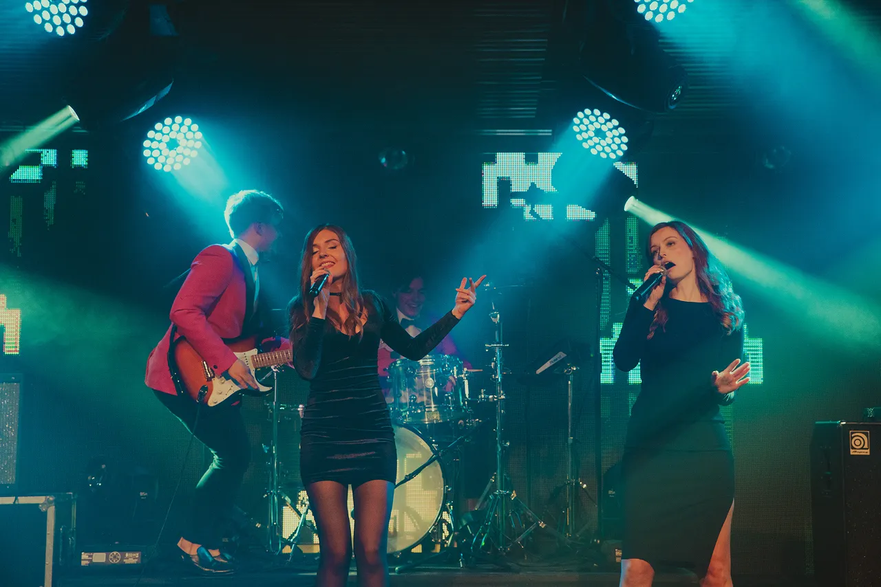 Four-piece band performing on stage with two female singers in black dresses, a guitarist in a red jacket, and a drummer under blue stage lights.