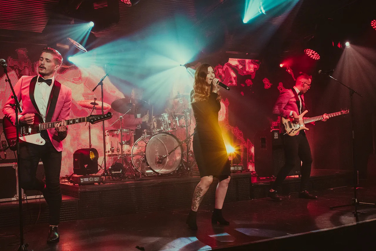 Band performing on stage with female vocalist holding a microphone and male musicians playing guitar and bass under colorful stage lights.